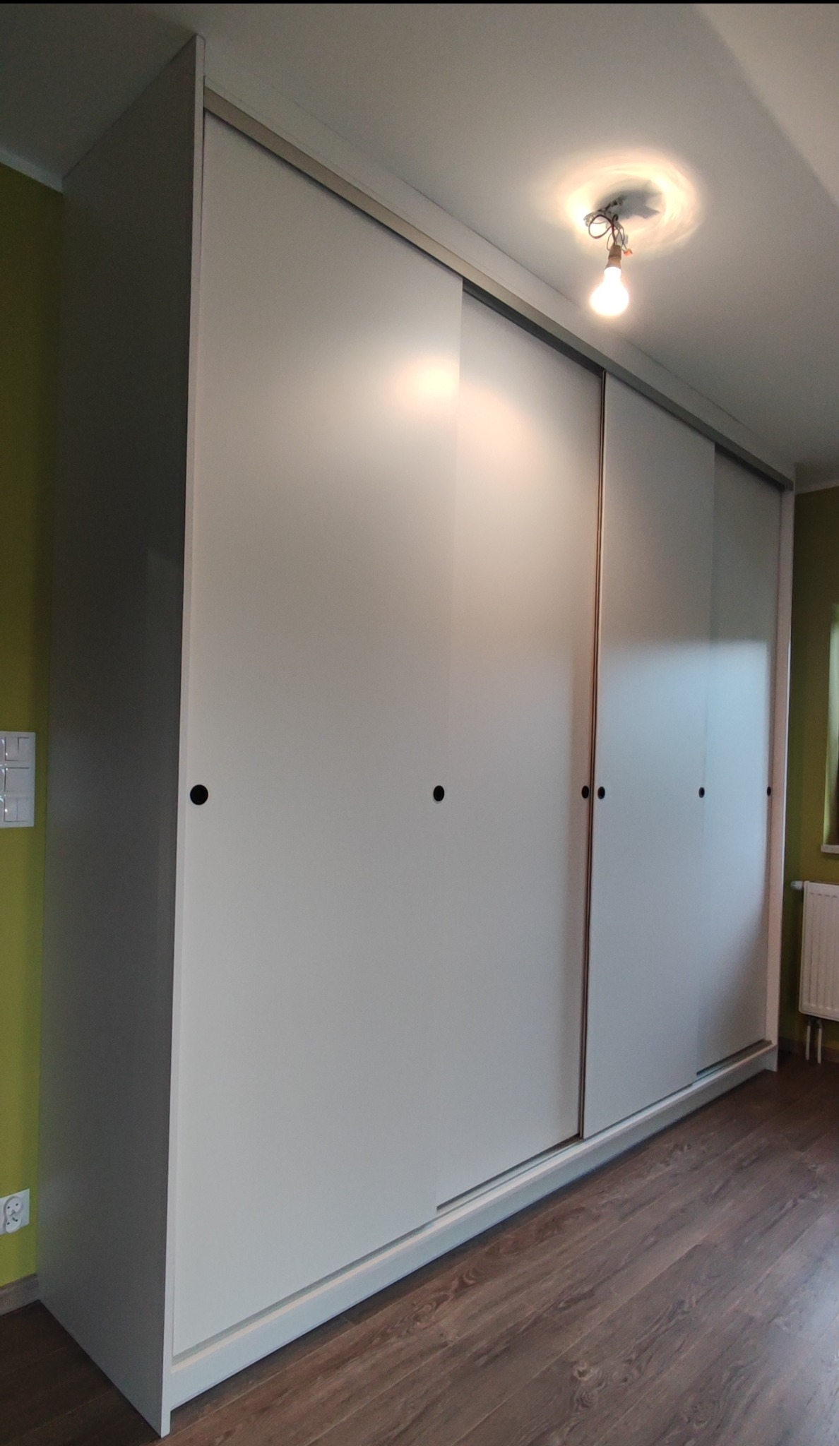 Szafa wnękowa z przesuwnymi, białymi drzwiami, widoczna na tle zielonej ściany i brązowej podłogi, oświetlona żarówką bez klosza