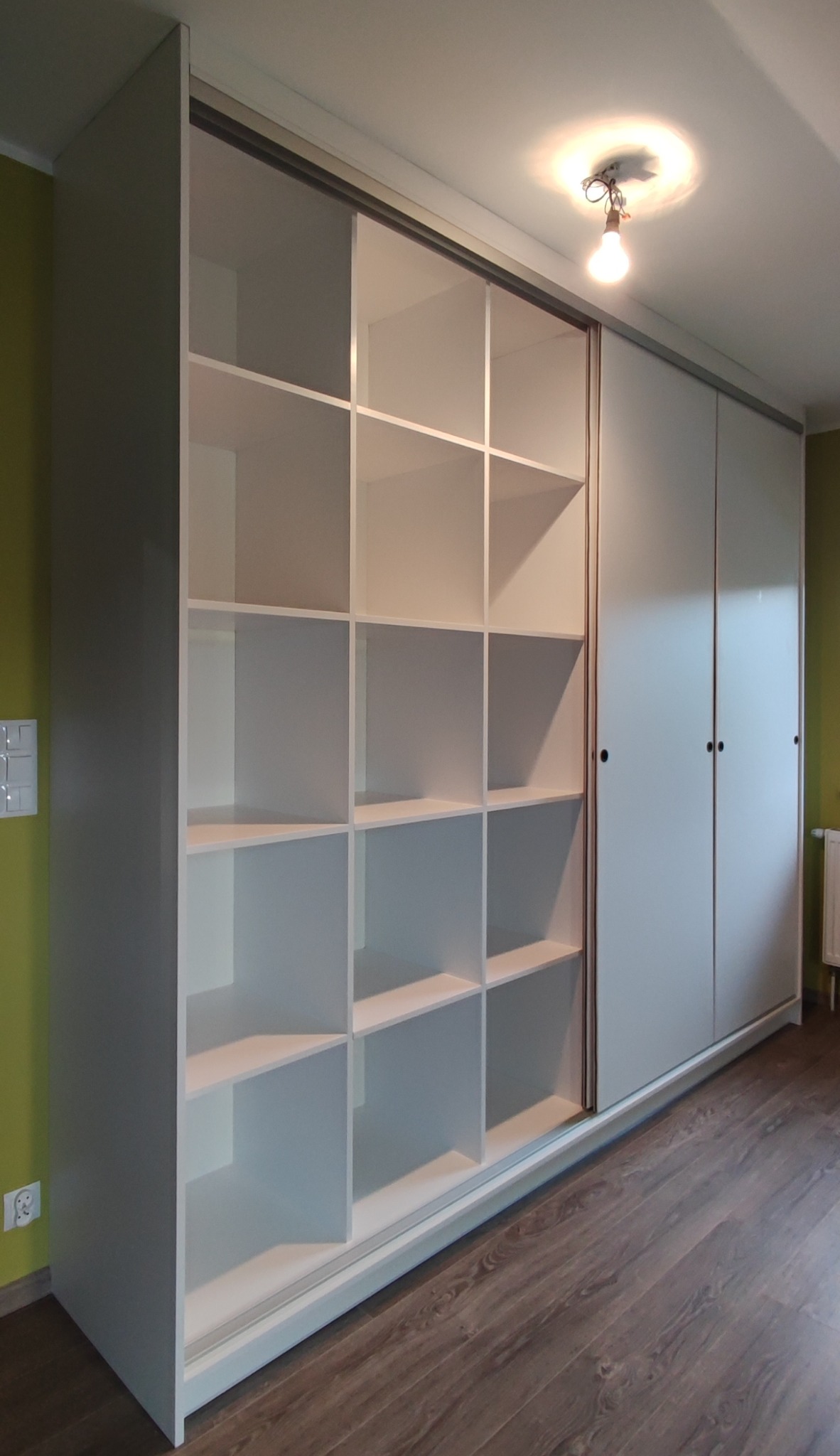 Biała szafa wnękowa z przesuwnymi drzwiami i otwartymi półkami, widoczna na tle zielonej ściany z włącznikiem i brązowej podłogi z paneli.