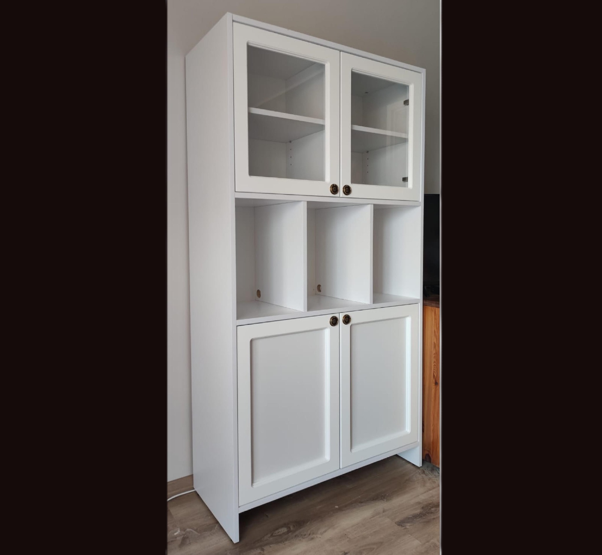 Biały regał z przeszklonymi drzwiczkami na górze i pełnymi drzwiczkami na dole, z czterema otwartymi półkami pośrodku, na tle ciemnej ściany i jasnej podłogi.