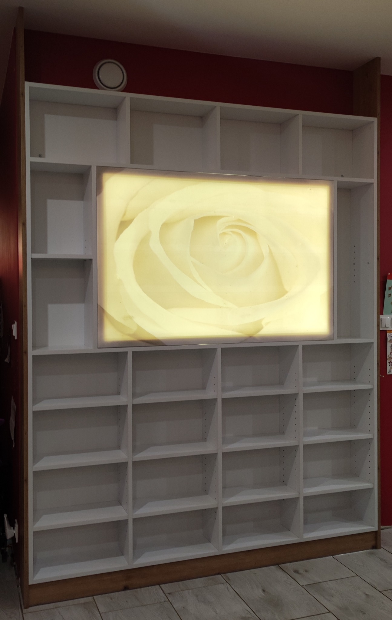 Biały regał z podświetlanym panelem z motywem róży, wbudowany w ścianę o czerwonym kolorze, widoczne rzędy półek.