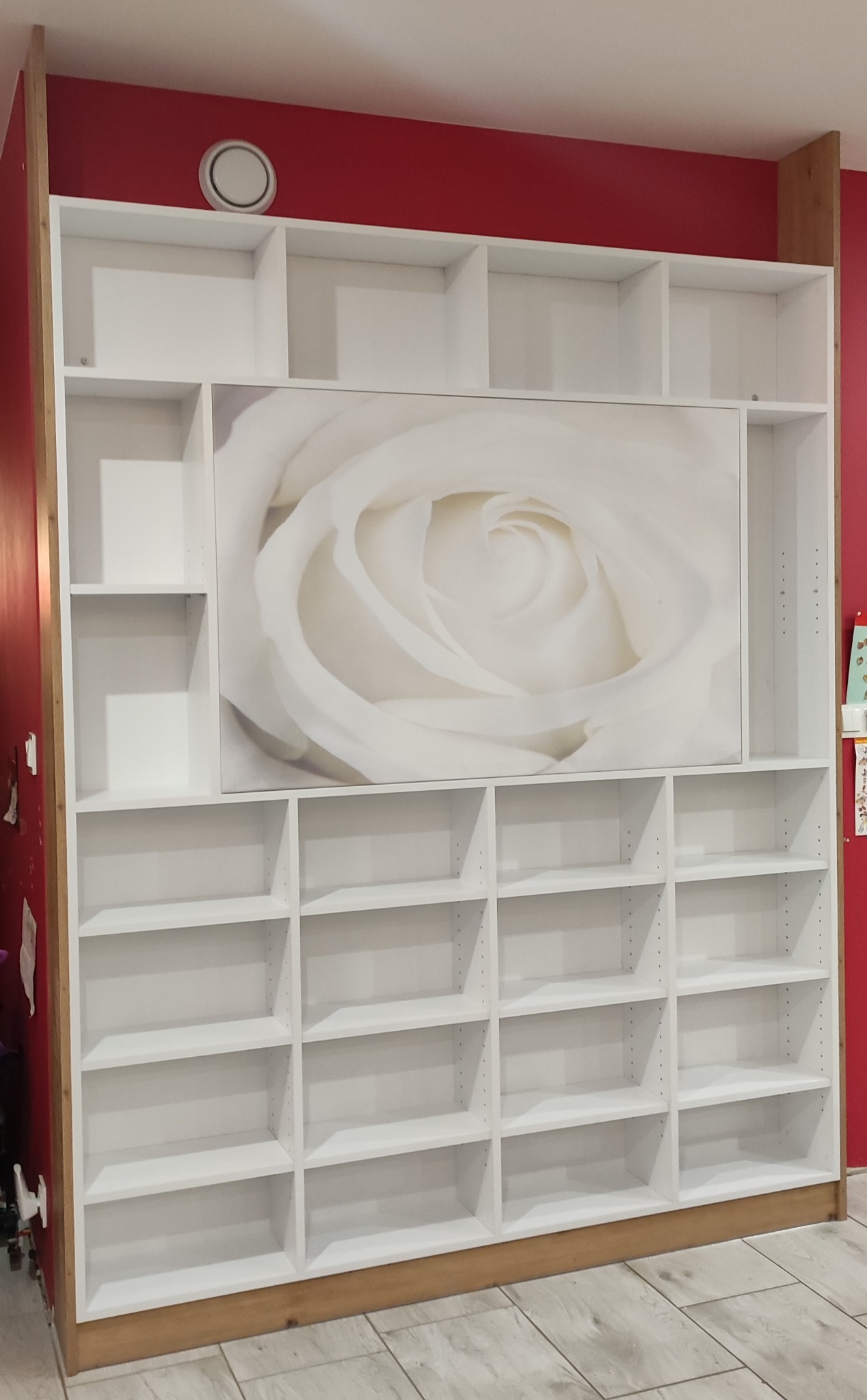 Biała, zabudowana szafa na wymiar z licznymi półkami, z centralnie umieszczonym panelem z grafiką białej róży, na tle czerwonej ściany.