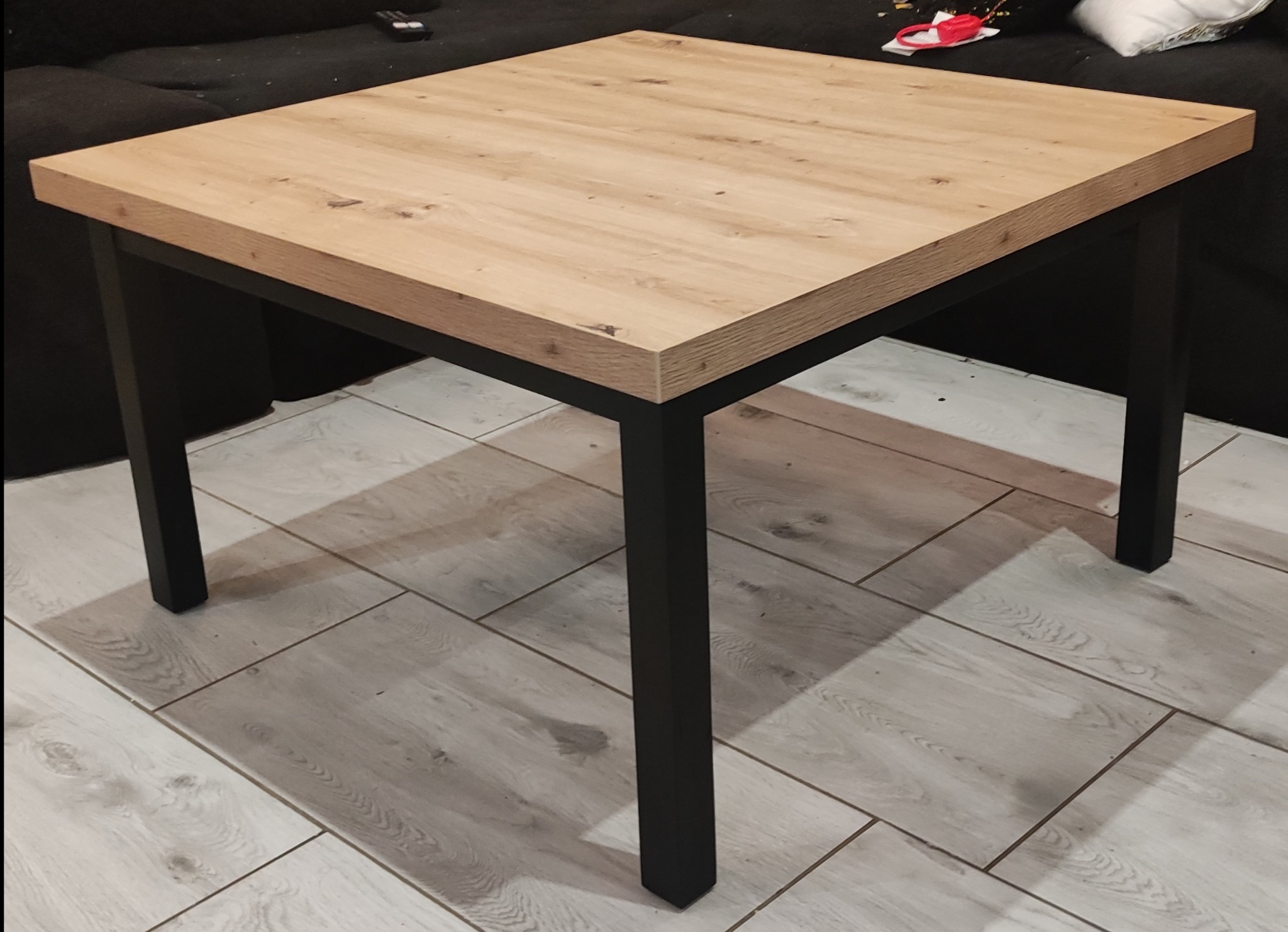 Kwadratowy stolik kawowy z blatem imitującym jasne drewno i czarnymi, metalowymi nogami, ustawiony na podłodze wyłożonej panelami imitującymi drewno.