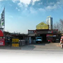 Szeroki kadr na skład budowlany z ekspozycją dachówek, blach i rynien. Widoczne banery reklamowe producentów pokryć dachowych i budynek biura z szyldem AS.