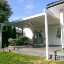 Biała pergola z aluminiową konstrukcją, zacieniająca taras z meblami ogrodowymi, przylegająca do jasnego domu z oknami i drzwiami tarasowymi, widok z zadbanego trawnika w słoneczny dzień.