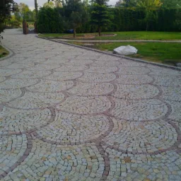 Oryginalna nawierzchnia z kostki granitowej ułożona w dekoracyjny wzór wachlarza, prowadząca do bramy w ogrodzie. Estetyczne wykończenie ścieżki.