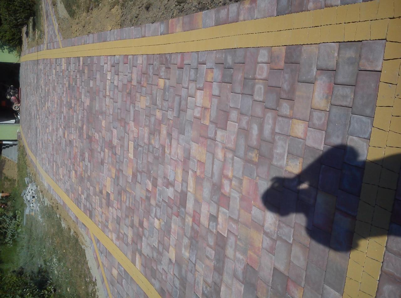 Ścieżka z kostki brukowej w odcieniach czerwieni i szarości, obrzeża wykończone żółtą kostką. Widoczny cień osoby. Ułożona kostka prowadzi wzdłuż zieleni.