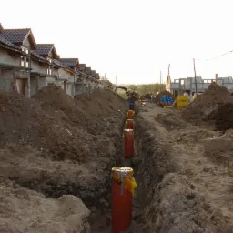 Wykop z pomarańczowymi rurami zabezpieczonymi ż&oacute;łtą folią, widoczne fundamenty budynk&oacute;w w tle, teren budowy.