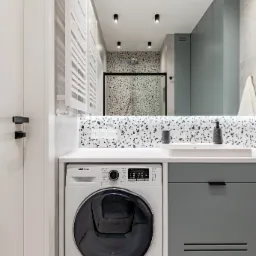 Nowoczesna łazienka z pralką Samsung pod umywalką, lustrem z oświetleniem punktowym i kabiną prysznicową w tle z mozaiką lastryko.