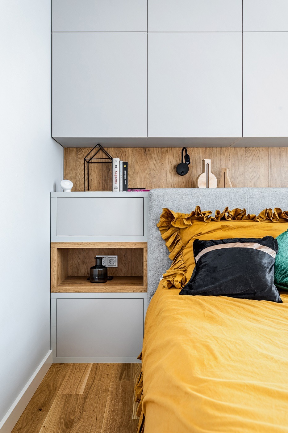 Nowoczesna sypialnia w Warszawie z zabudową meblową nad łóżkiem, drewnianym panelem i żółtą pościelą z falbanami, uzupełniona czarną lampką nocną i dekoracyjnymi przedmiotami.