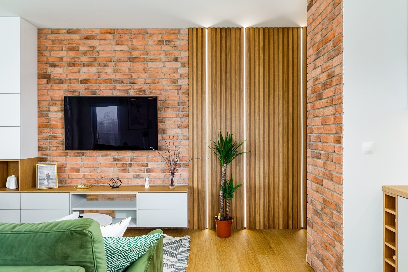 Nowoczesny salon z ceglaną ścianą, telewizorem, drewnianymi panelami z oświetleniem LED, białą szafką RTV, zieloną sofą i rośliną doniczkową.