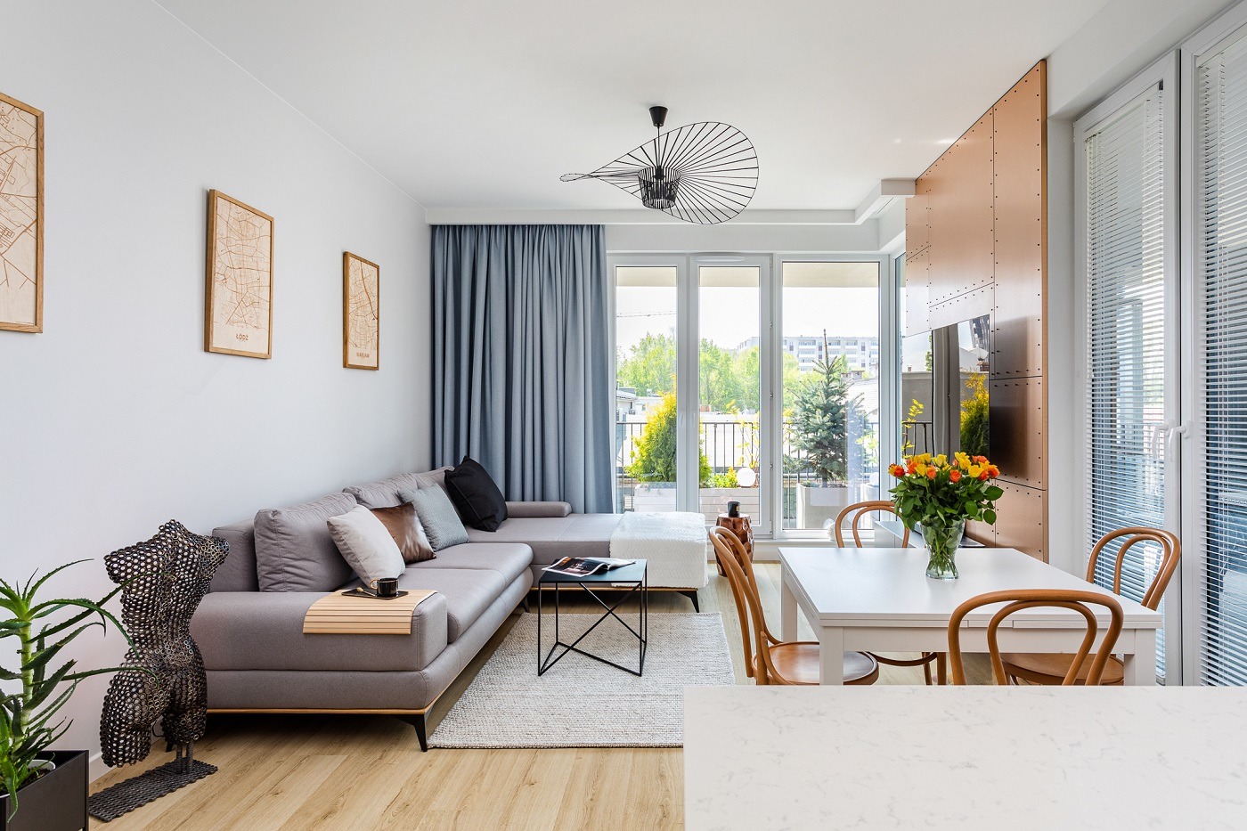 Jasne, nowoczesne wnętrze salonu z szarą sofą narożną, drewnianymi mapami na ścianie, geometryczną lampą sufitową i stołem jadalnym z bukietem różnokolorowych róż.