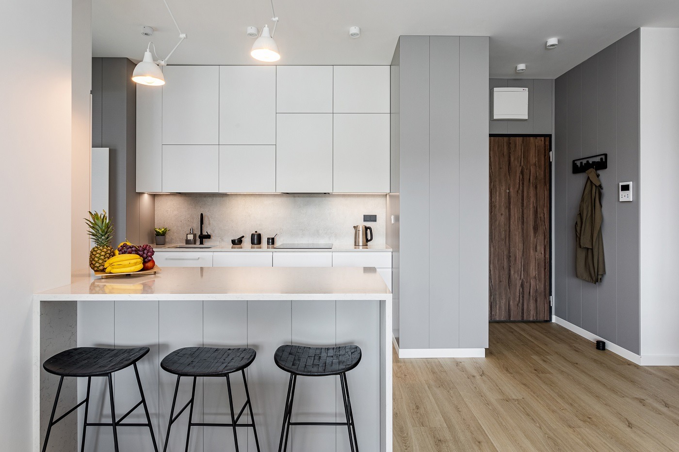 Jasna kuchnia w minimalistycznym stylu z wyspą, owocami na blacie i trzema czarnymi hokerami, widok na przedpokój z drewnianymi drzwiami i wieszakiem na ubrania.