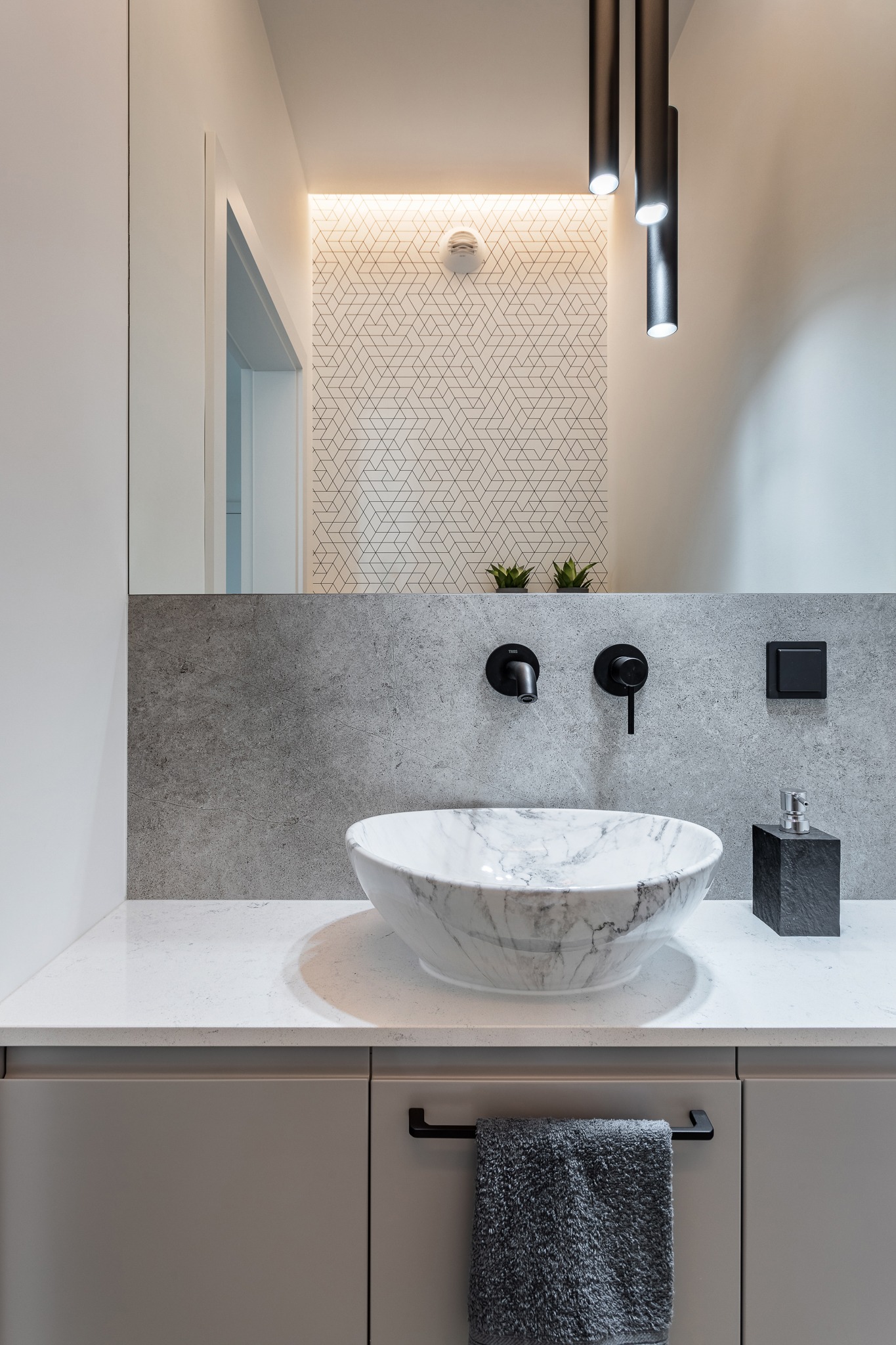 Nowoczesna łazienka z umywalką w kształcie misy z marmuru, czarną armaturą, szarymi płytkami i geometryczną tapetą w tle, oświetlona trzema cylindrycznymi lampami sufitowymi.
