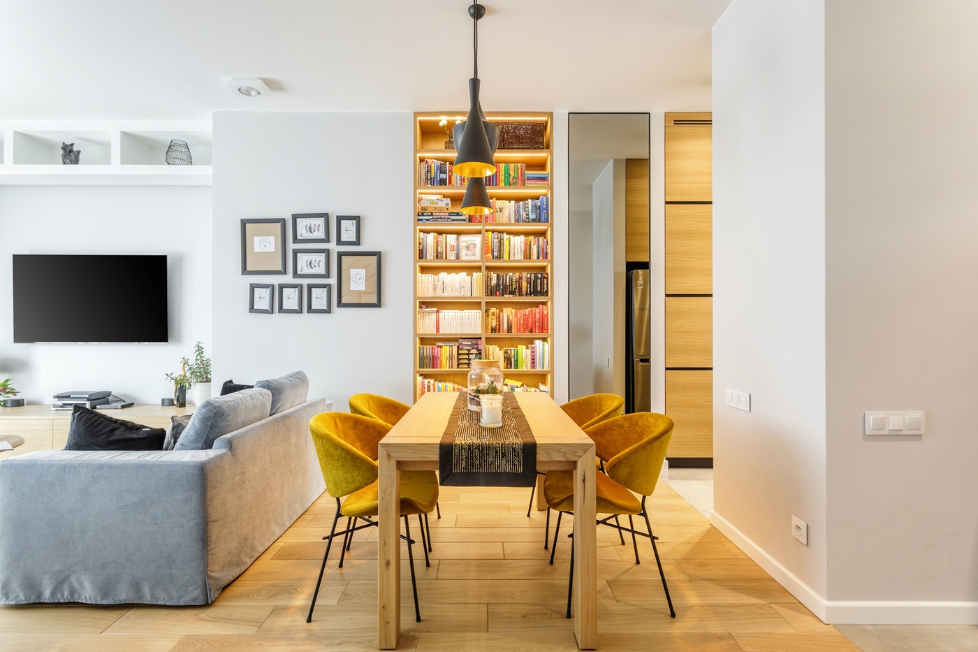 Jasne wnętrze salonu z drewnianym stołem, żółtymi krzesłami, szarą sofą i regałem z książkami podświetlonym od wewnątrz.