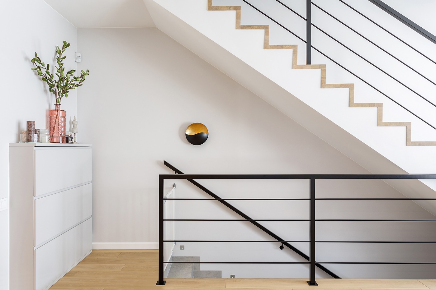 Nowoczesne wnętrze: biała szafka na buty z dekoracjami, schody z czarną balustradą i okrągła dekoracja ścienna w kolorach złota i czerni.
