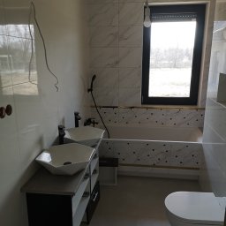 Marcin Plewak Lolek P.H.U. - Wnętrze łazienki z dwoma umywalkami nablatowymi na szafce, białą wanną obudowaną płytkami z geometrycznym wzorem, wiszącą toaletą i dużym oknem z roletami; widoczne niedokończone podłączenia...