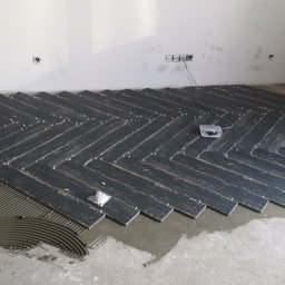 Marcin Plewak Lolek P.H.U. - Układanie ciemnych płytek podłogowych w jodełkę, widoczne krzyżyki dystansowe i rozprowadzony klej, fragment ściany z gniazdkami elektrycznymi.