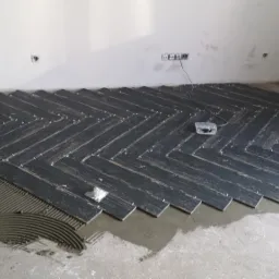 Układanie ciemnych płytek podłogowych w jodełkę, widoczne krzyżyki dystansowe i rozprowadzony klej, fragment ściany z gniazdkami elektrycznymi.