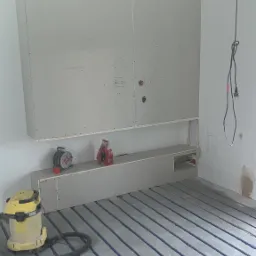 Montaż płyty karton gips na ścianie z wnęką, widoczne rury ogrzewania podłogowego, odkurzacz przemysłowy i zwinięty przedłużacz na półce w trakcie prac wykończeniowych.