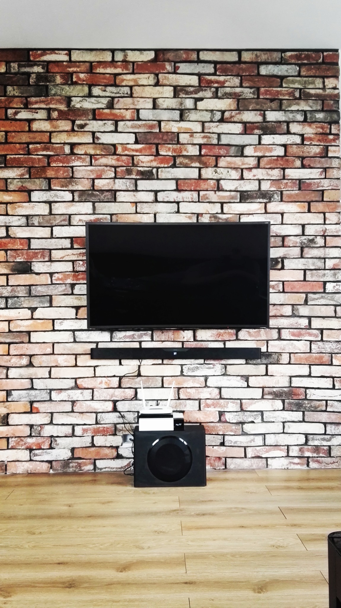 Telewizor zawieszony na dekoracyjnej ścianie z cegły w salonie, z soundbarem i subwooferem poniżej.