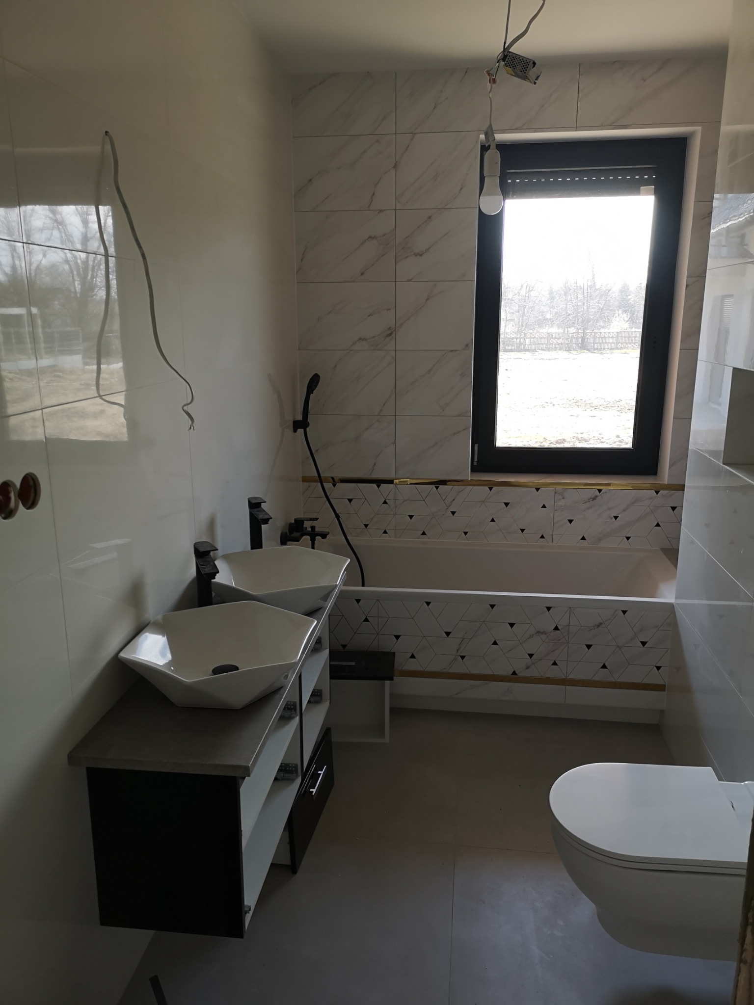 Wnętrze łazienki z dwoma umywalkami nablatowymi na szafce, białą wanną obudowaną płytkami z geometrycznym wzorem, wiszącą toaletą i dużym oknem z roletami; widoczne niedokończone podłączenia...