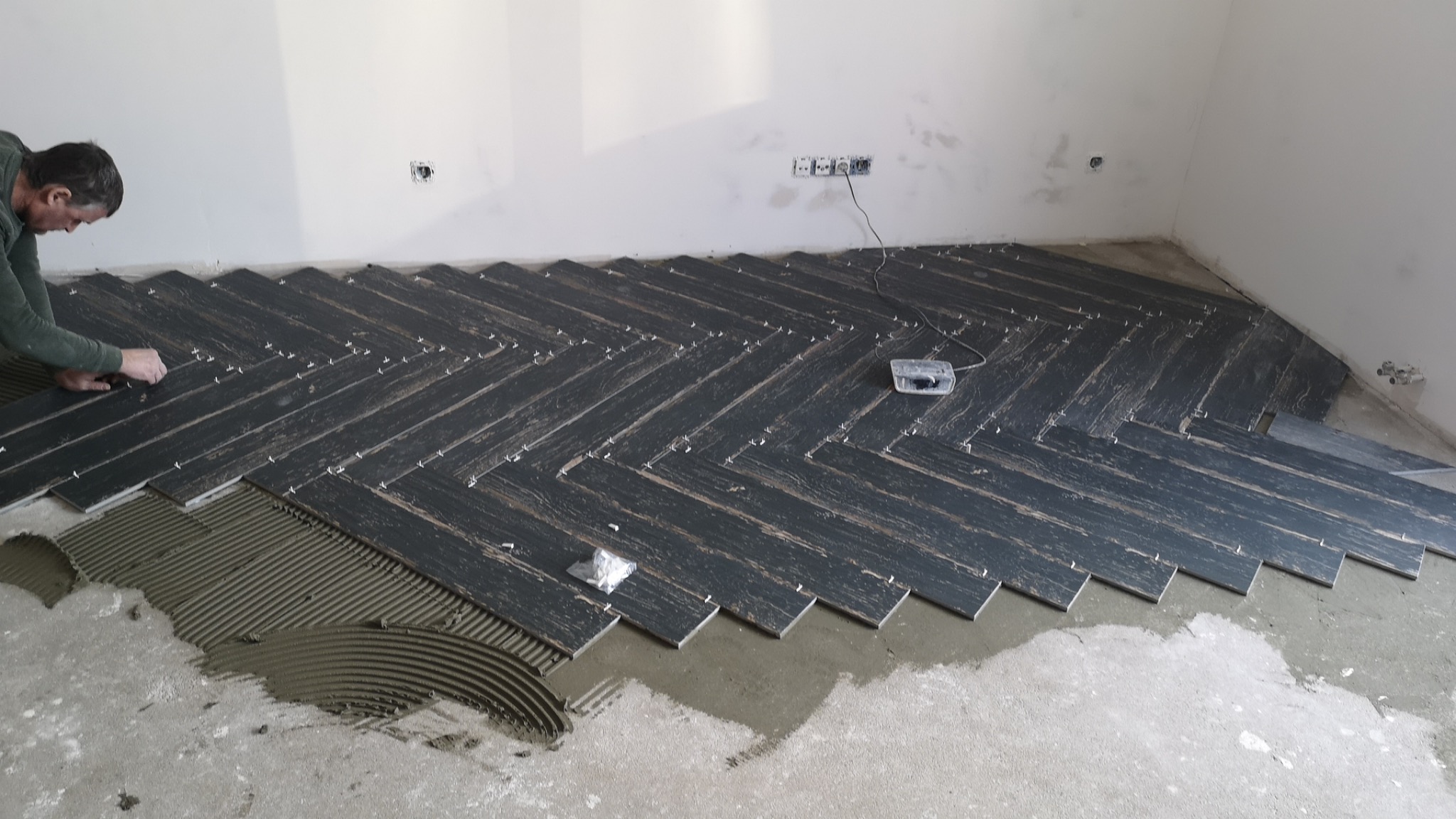 Układanie ciemnych płytek podłogowych w jodełkę, widoczne krzyżyki dystansowe i rozprowadzony klej, fragment ściany z gniazdkami elektrycznymi.