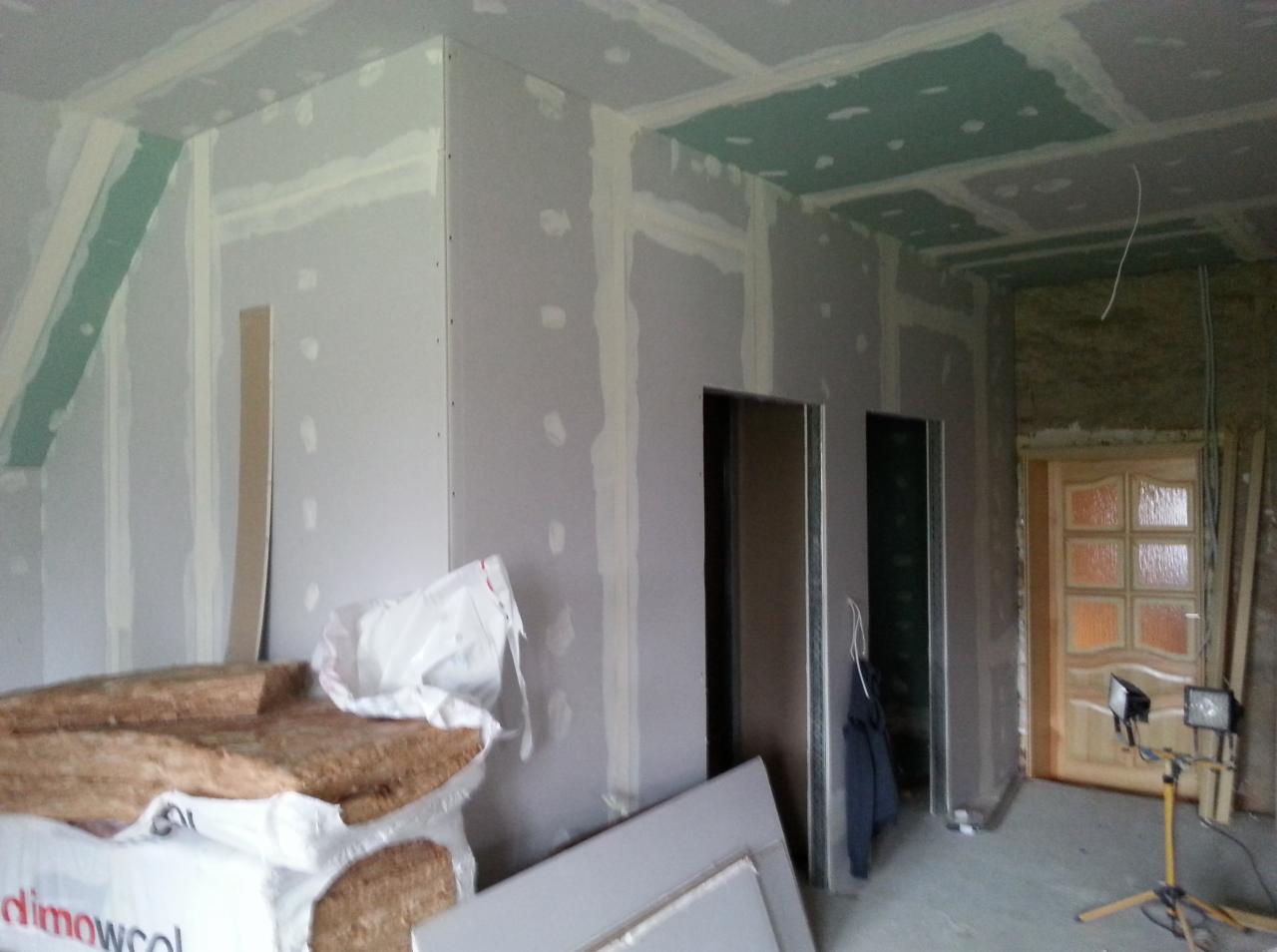 Wnętrze w trakcie montażu płyt karton-gips, widoczne ocieplenie, szpachlowane łączenia, przygotowane otwory na drzwi i oświetlenie budowlane.