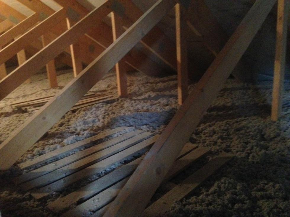 Poddasze z drewnianą konstrukcją dachu i rozłożoną warstwą izolacji z wełny mineralnej, widoczne deski podłogowe do ułożenia, słabe oświetlenie.