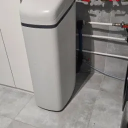 Biały zmiękczacz wody marki Viessmann Aquahome 25, zainstalowany w pomieszczeniu z szarymi płytkami, widoczne przyłącza hydrauliczne.
