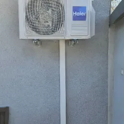 Biała jednostka zewnętrzna klimatyzatora Haier zamontowana na szarej ścianie z widocznymi elementami instalacji.