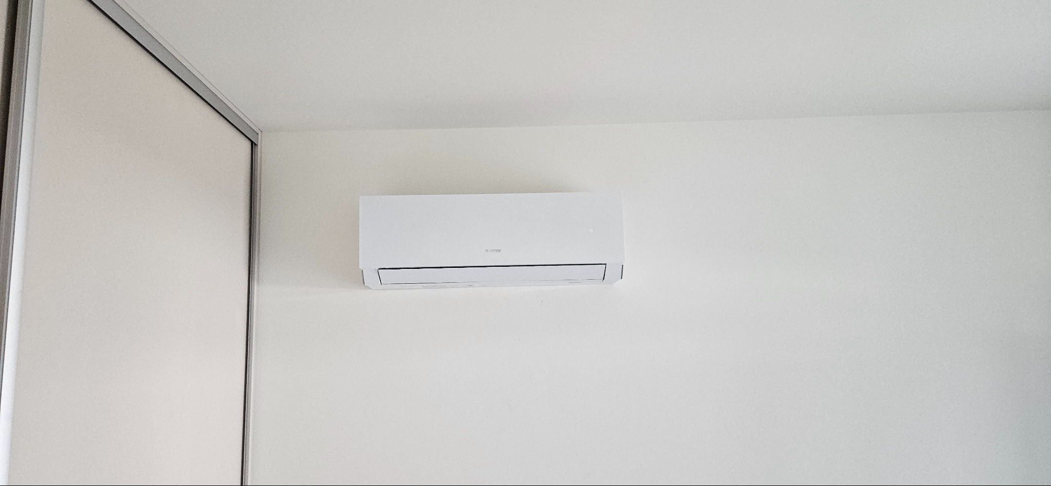 Biała jednostka wewnętrzna klimatyzacji zamontowana na jasnej ścianie w minimalistycznym wnętrzu, widoczny fragment szafy przesuwnej po lewej stronie.
