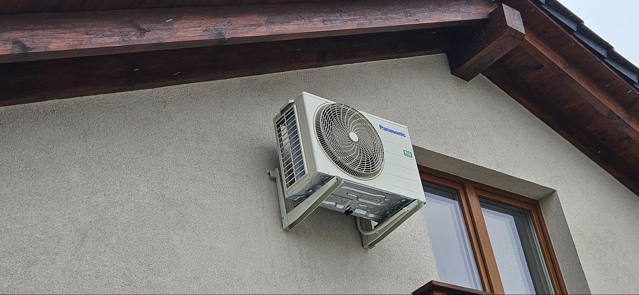 Zewnętrzna jednostka klimatyzacji Panasonic zamontowana na elewacji domu obok okna z drewnianą ramą, widoczne brązowe elementy konstrukcji dachu.