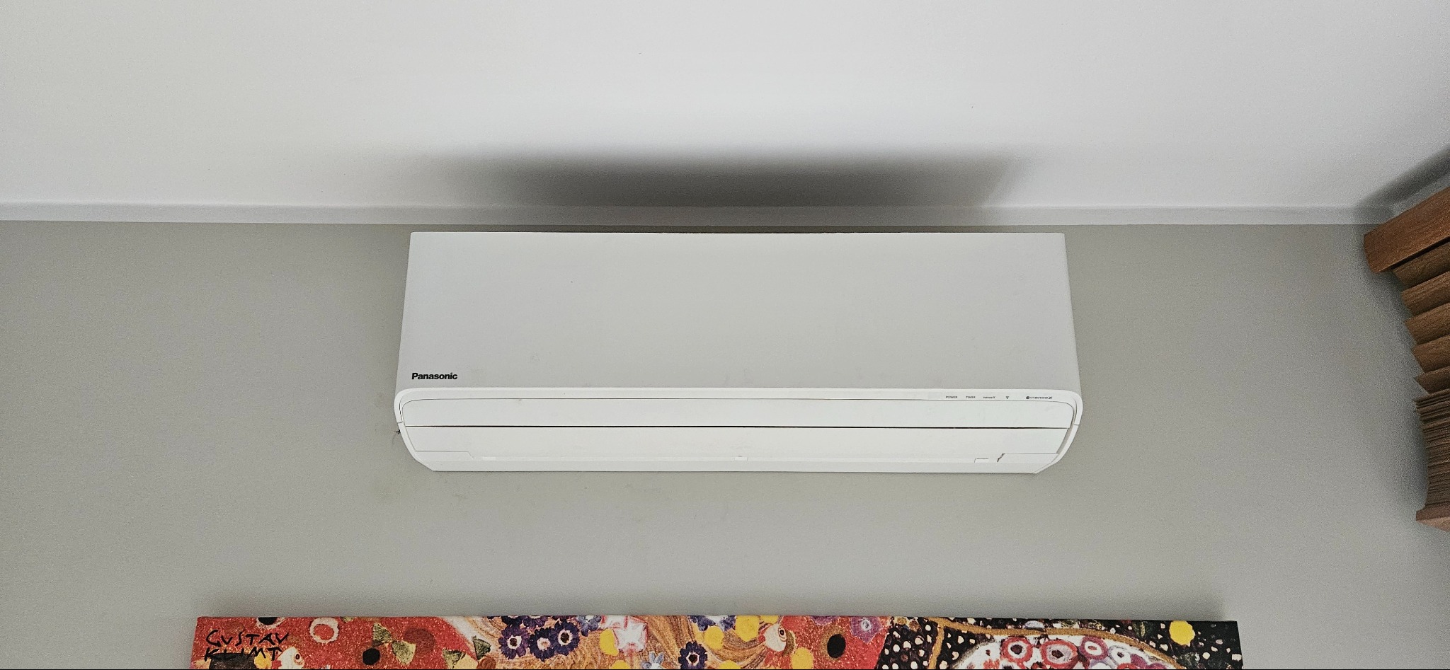 Biały klimatyzator Panasonic zamontowany na szarej ścianie w domu, widoczne elementy sterujące i obrazek w stylu Gustava Klimta poniżej.