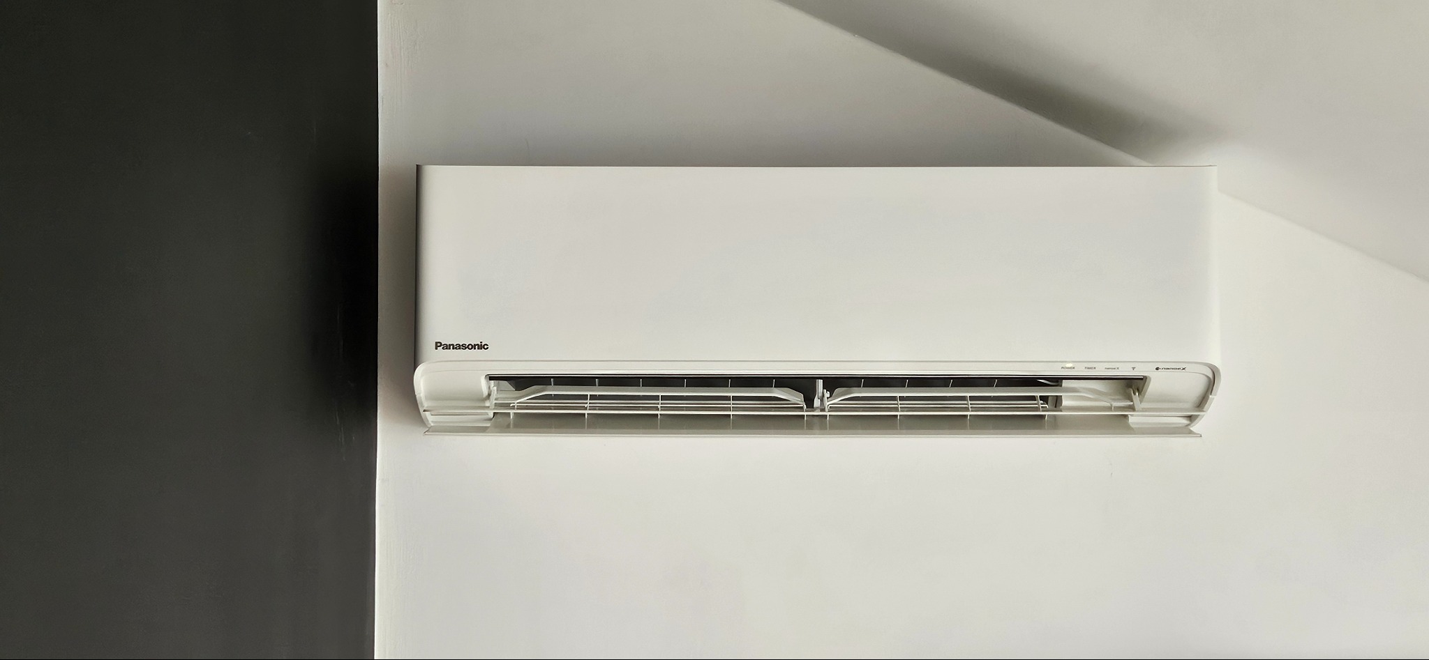Biała jednostka klimatyzacji Panasonic zamontowana na białej ścianie, z fragmentem czarnej ściany po lewej stronie. Widoczne szczeliny wylotu powietrza i subtelne cienie na ścianie.