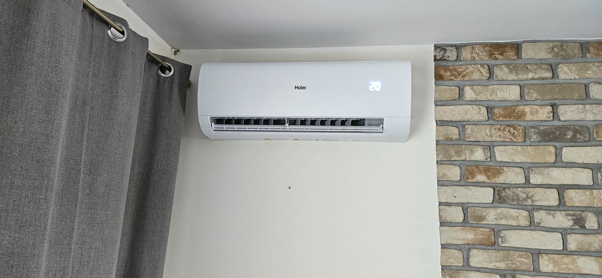 Biały klimatyzator Haier z wyświetlaczem ustawionym na 20 stopni Celsjusza, zamontowany na ścianie w Ceradzu Dolnym, obok fragmentu dekoracyjnej ściany z cegły i zasłony w tle.