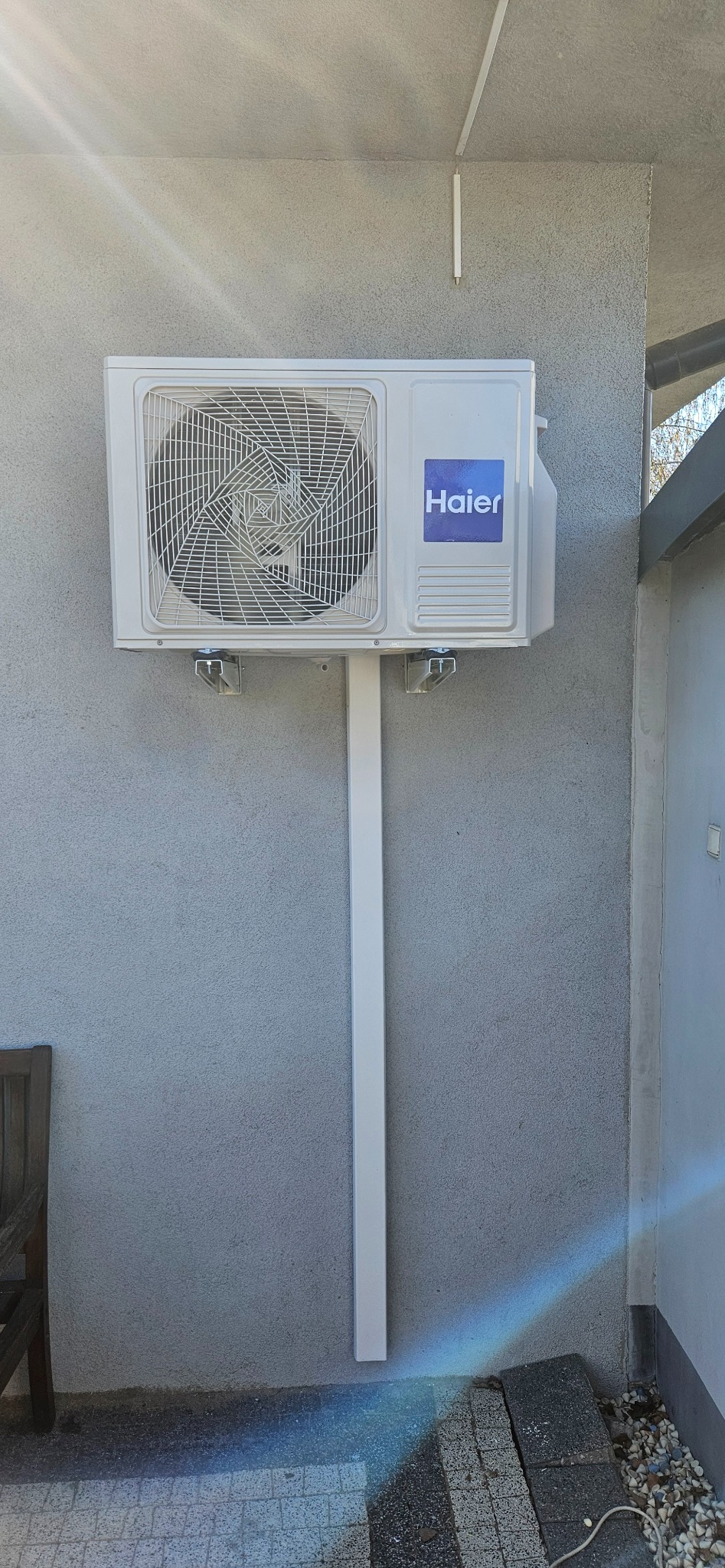 Biała jednostka zewnętrzna klimatyzatora Haier zamontowana na szarej ścianie z widocznymi elementami instalacji.