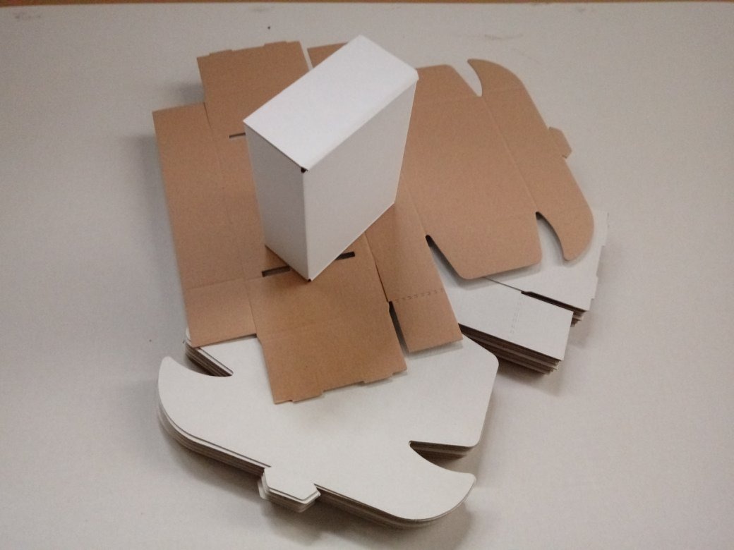 Białe pudełko kartonowe złożone, otoczone płaskimi, wyciętymi elementami z brązowego i białego kartonu, gotowymi do złożenia.