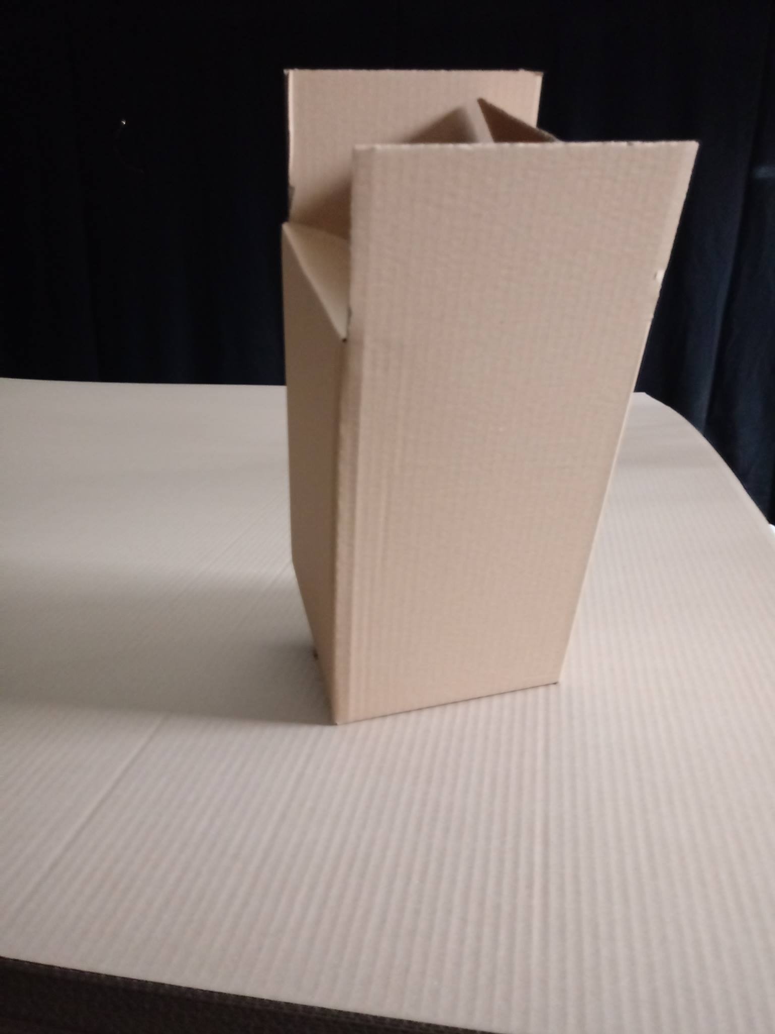 Nierozłożone pudełko z tektury falistej w kształcie trapezu, widoczne klapy górne, ustawione na powierzchni z tektury falistej, czarne tło.