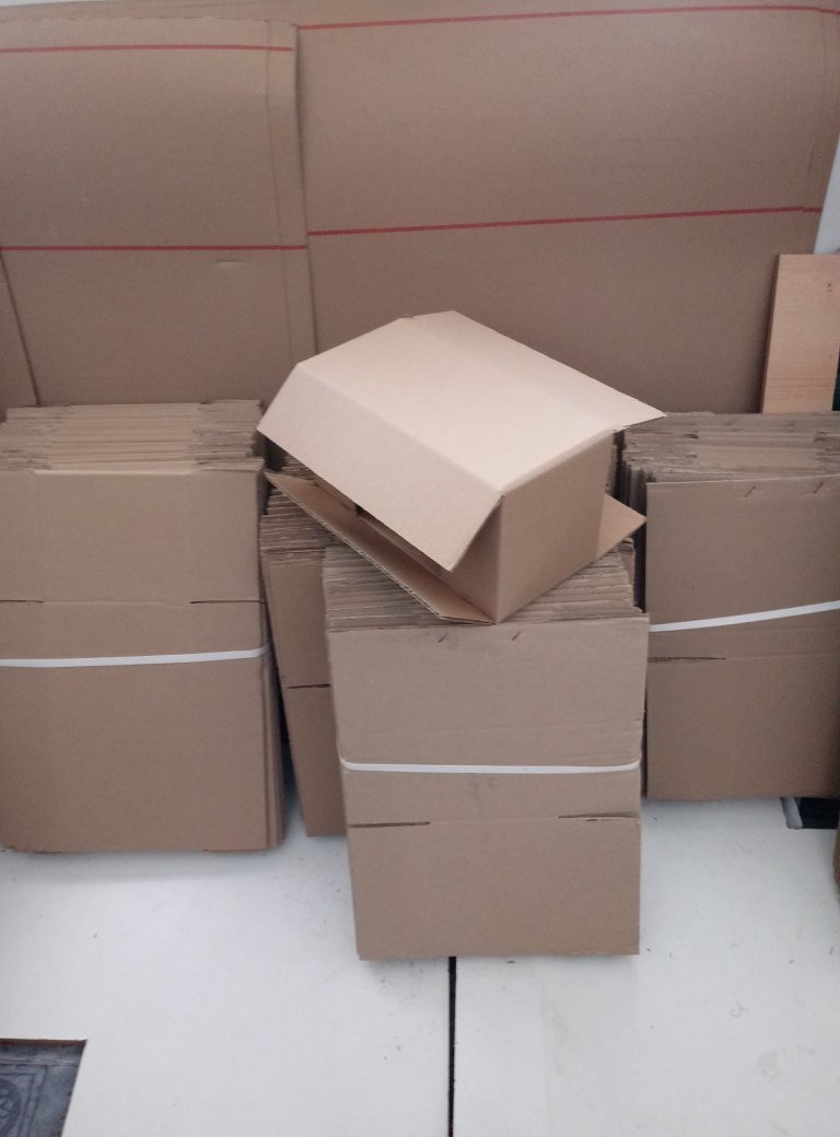 Stosy płaskich kartonowych pudełek, zabezpieczonych białym sznurkiem, z jednym rozłożonym pudełkiem na wierzchu, na tle dużych arkuszy kartonu z czerwonymi liniami.