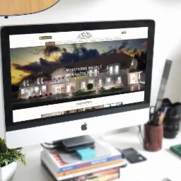 Ekran komputera iMac wyświetlający wizualizację architektoniczną budynku z napisem 'Wyjątkowe miejsce na każdą okazję', na biurku obok roślina doniczkowa, drewniana figurka i przybory biurowe.