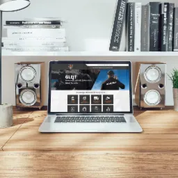 Laptop wyświetlający stronę internetową Agencji Ochrony Glejt na drewnianym biurku, obok głośniki i lampka, na półce książki i roślina w metalowej doniczce.