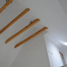 Surowe drewniane belki stropowe na tle białego sufitu poddasza, widoczne kable elektryczne i okno dachowe w trakcie montażu.