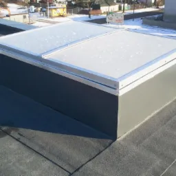 Świetlik dachowy z aluminiową ramą na pokryciu bitumicznym dachu płaskiego, widok z góry na tle zabudowań miejskich w zimowej scenerii.