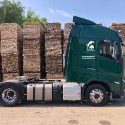 Zielony samochód ciężarowy firmy DREWNEX z logo, stojący na tle stosów pociętego drewna.