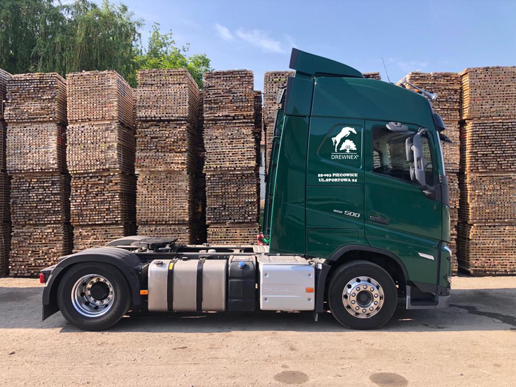Zielony samochód ciężarowy firmy DREWNEX z logo, stojący na tle stosów pociętego drewna.