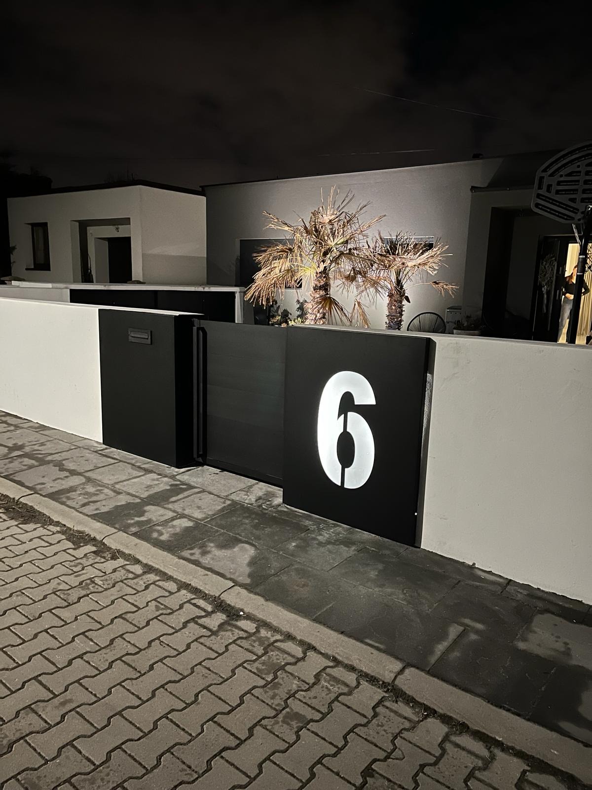 Nowoczesna, czarna brama posesji z podświetlonym numerem '6' w białym kolorze, widoczna nocą na tle białego muru i domu z palmami.