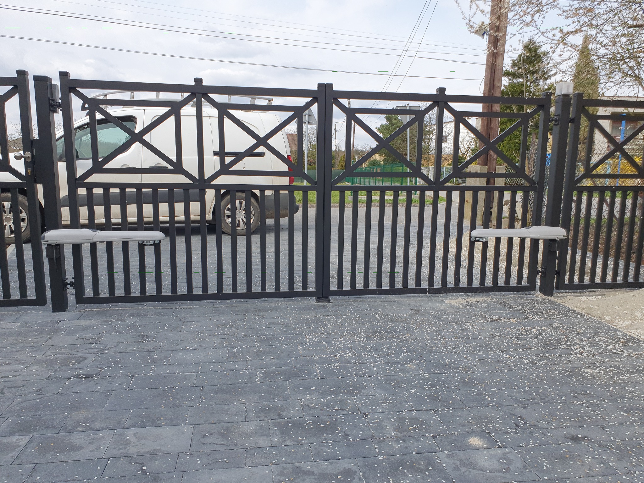 Automatyczna brama dwuskrzydłowa, czarna, ze wzorem krzyży, z napędem. Na podjeździe leżą opadłe płatki.