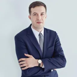 Portret mężczyzny w granatowym garniturze, białej koszuli i szarym krawacie, z zegarkiem na lewym nadgarstku, stojącego na jasnym tle z założonymi rękami.