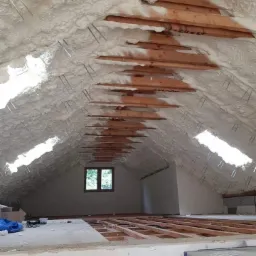 Poddasze z drewnianą konstrukcją dachu pokryte warstwą białej piany izolacyjnej, widoczne metalowe mocowania i fragment okna dachowego.