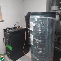 Kotłownia z nowym, owiniętym folią zbiornikiem akumulacyjnym Kospel SB-300 i podłączonym, używanym kotłem na paliwo stałe marki Viadrus, widoczne przewody i instalacja hydrauliczna